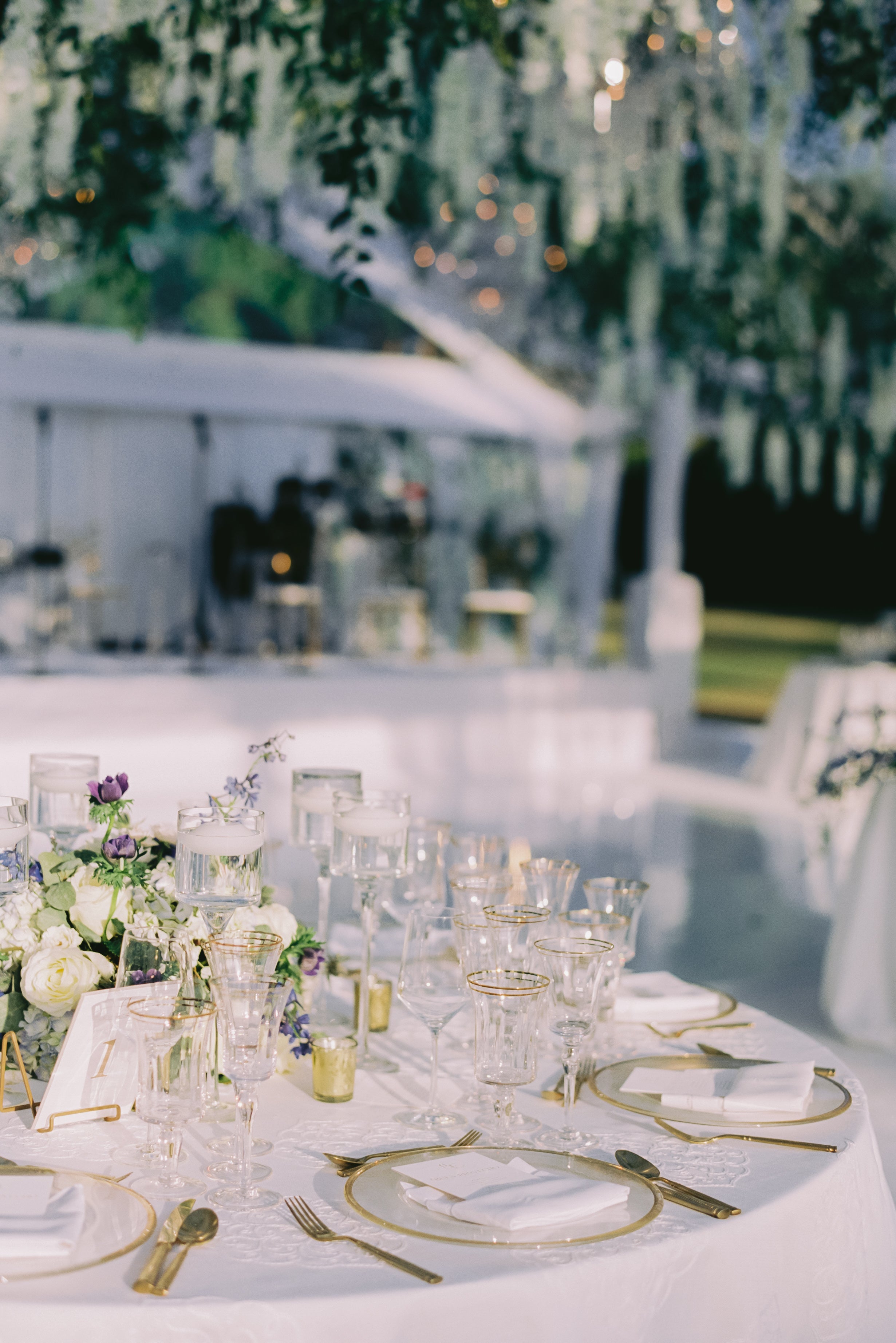 Elegant outdoor dining table setting with white tablecloth, gold cutlery, and floral arrangements.